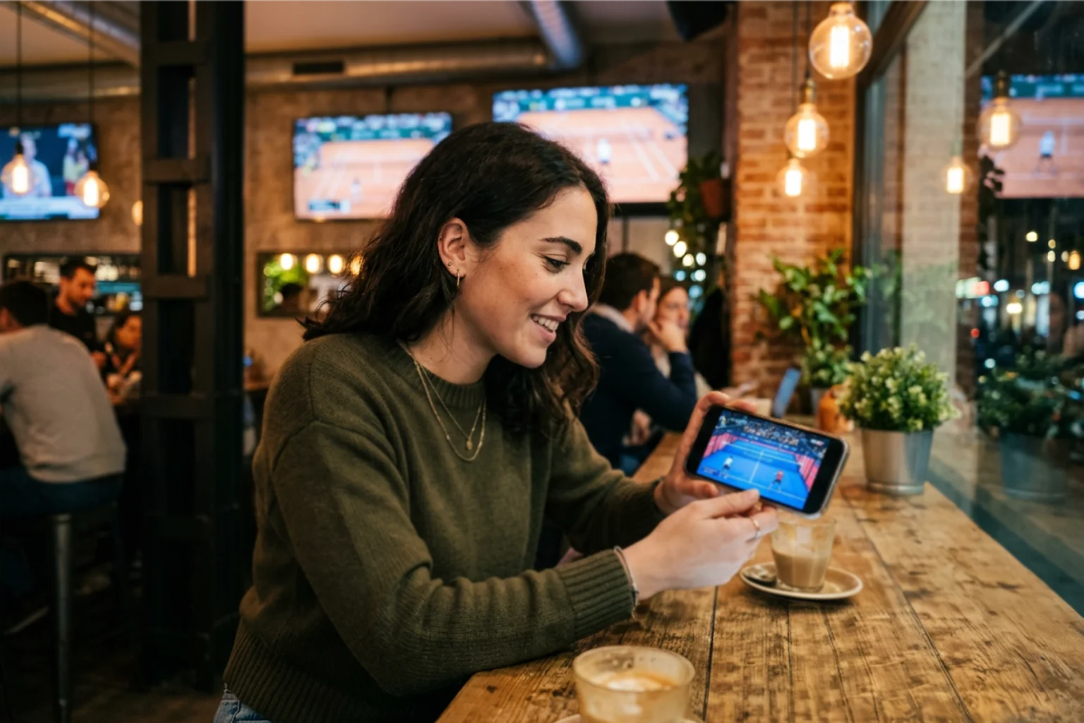 Mujer joven viendo partido de pádel en directo en su smartphone en un bar deportivo moderno con ambiente cálido